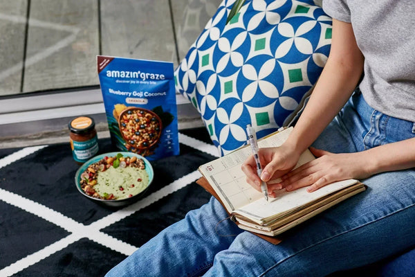 Woman writing in planner beside green smoothie bowl and Amazin’ Graze Blueberry Goji Coconut Granola pack.