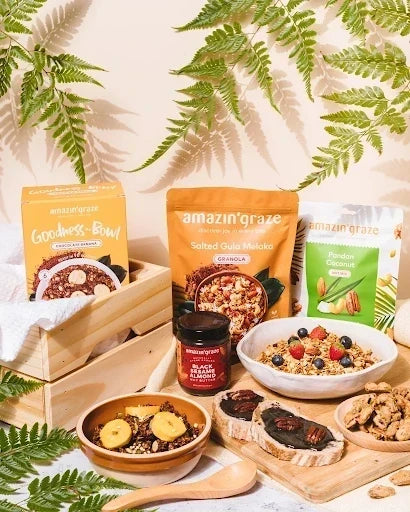Amazin’ Graze breakfast and snack products displayed with prepared dishes on wooden boards beneath green fern leaves.