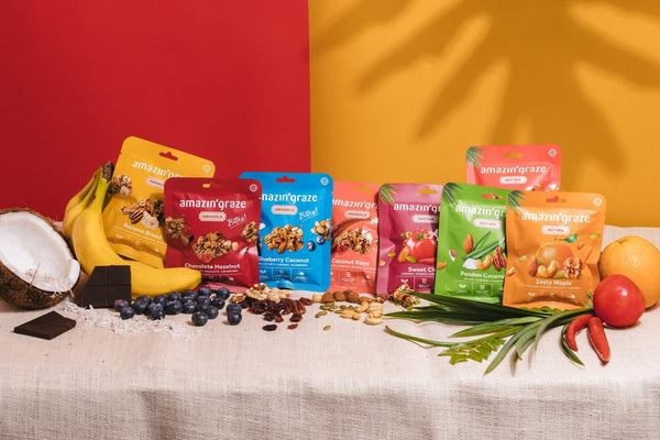 Amazin’ Graze granola bites and nut mixes displayed with fruits, coconut, and chocolate.