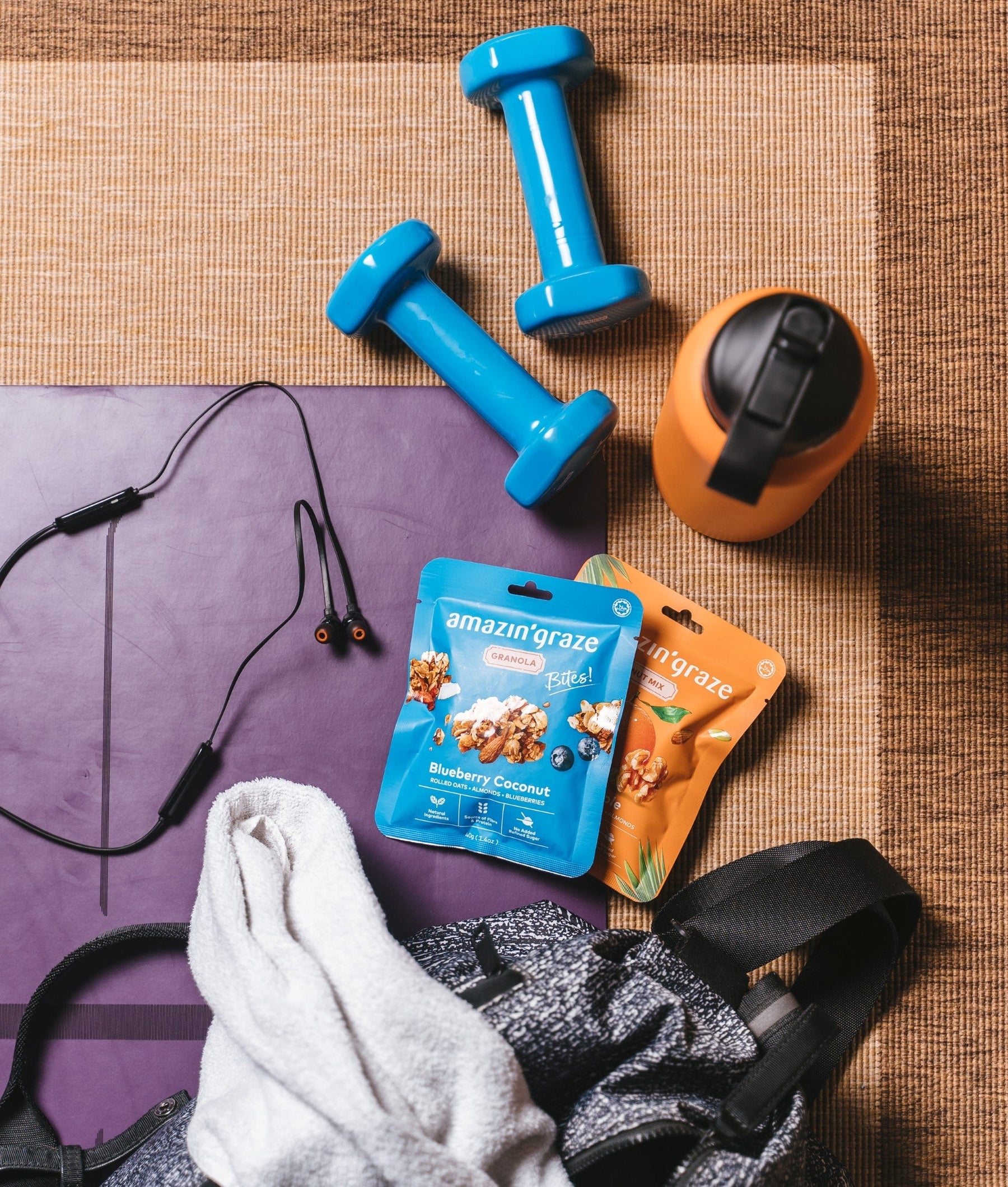 Amazin’ Graze plant-based snack packs beside workout gear, dumbbells, and a water bottle.