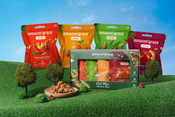 Amazin’ Graze nut mix variety box and snack packs displayed on a playful green landscape.