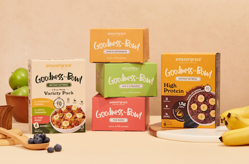 Amazin' Graze Goodness Bowl oatmeal boxes displayed with bananas, apples, blueberries, and serving bowls.